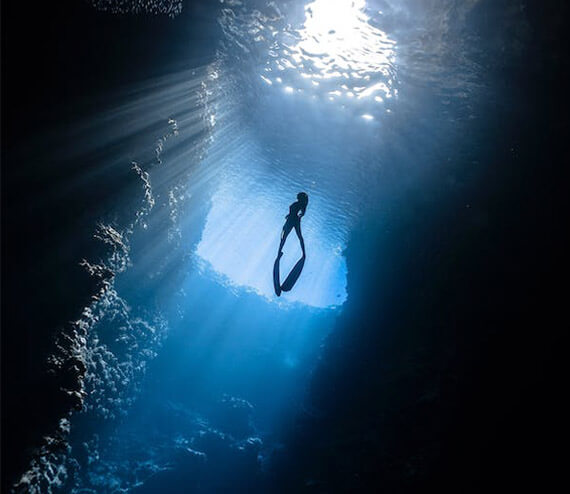 A scuba diver swimming towards the surface of the water and illuminated by light rays spilling in from above
