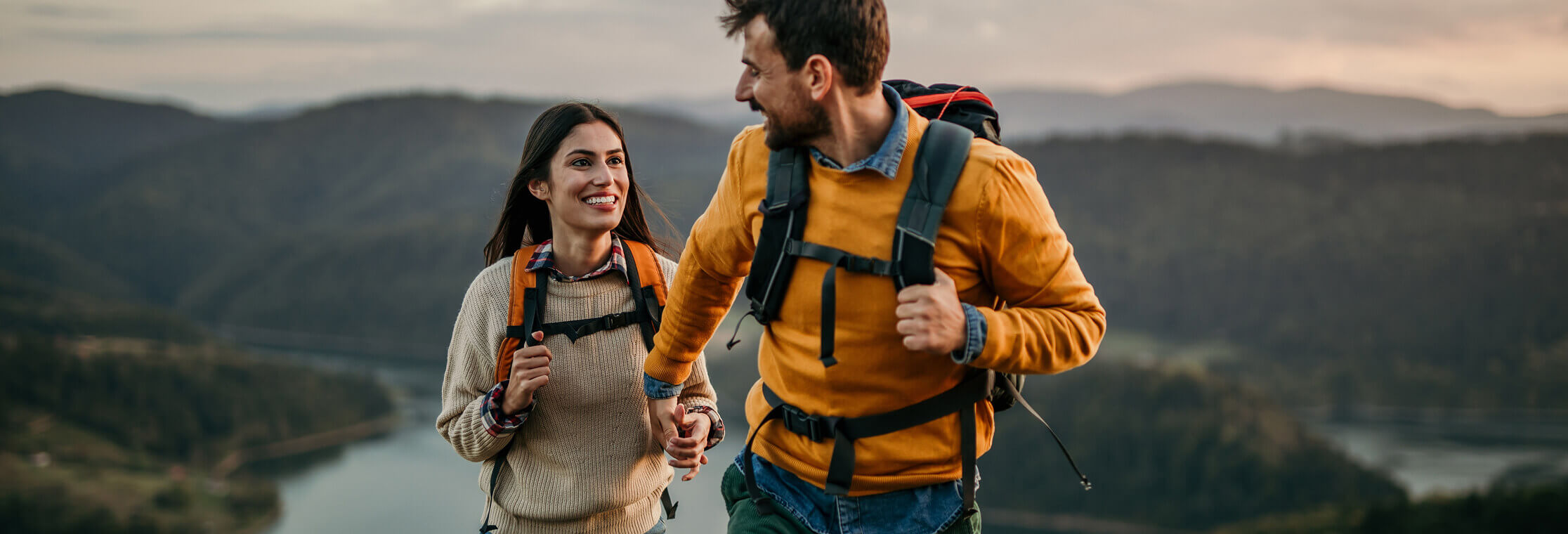 A man and woman are holding hands smiling at each other as they backpack up a mountain.