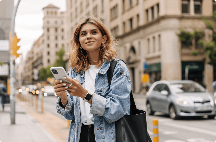 A young woman in a denim jacket looks up from her phone and smiles as she walks in the city center