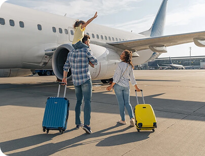 A family with a small child on dad's shoulders rolls their suitcases toward a jet plane for boarding