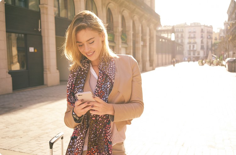 Young woman with a silk scarf and suitcase stops to check her phone in the middle of a European city