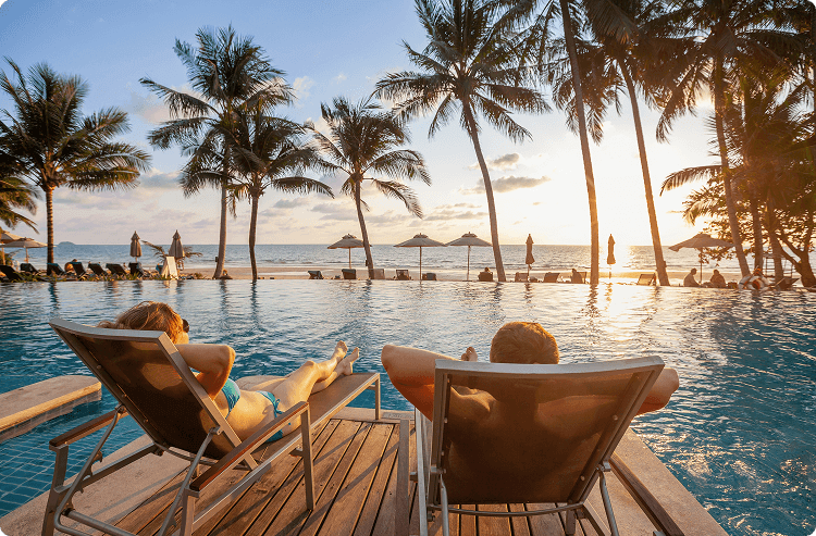 Couple relaxes on lounge chairs by an infinity pool at sunset, and watches the sunset over the ocean at an all-inclusive resort