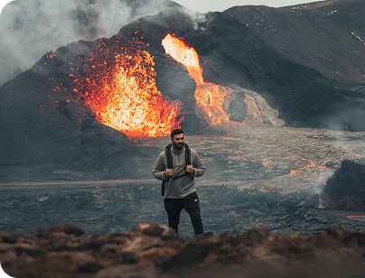 A traveler stands near an active volcano with lava erupting in the background