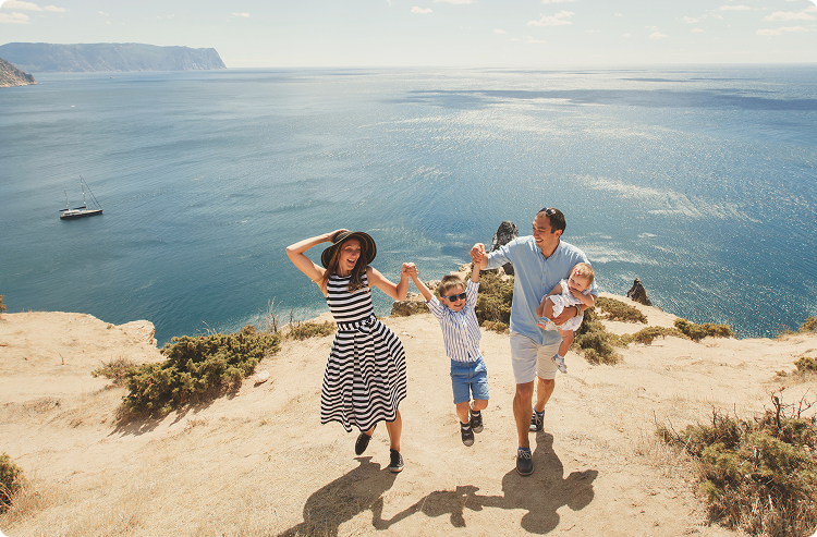 Family of four walking along a sunny cliffside overlooking the ocean; the parents hold hands with their son mid-jump, while the father carries a baby, with a sailboat and distant cliffs in the background