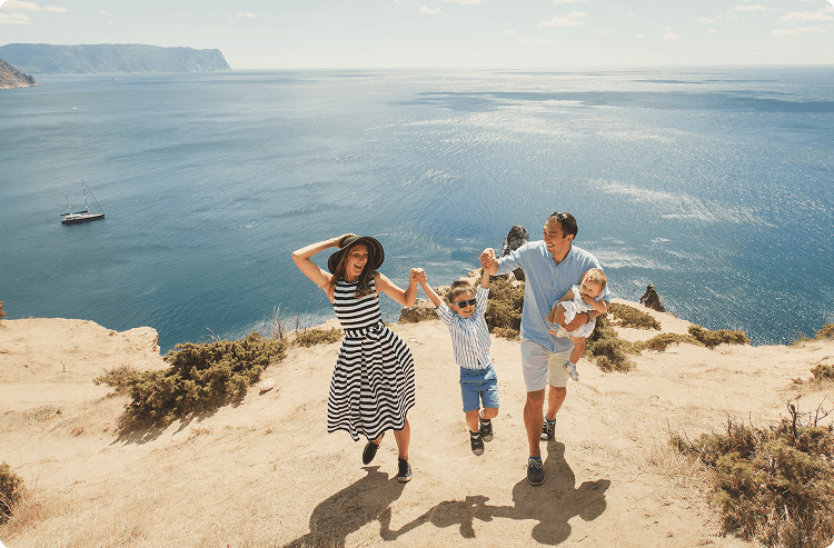 Family of four walking along a sunny cliffside overlooking the ocean; the parents hold hands with their son mid-jump, while the father carries a baby, with a sailboat and distant cliffs in the background