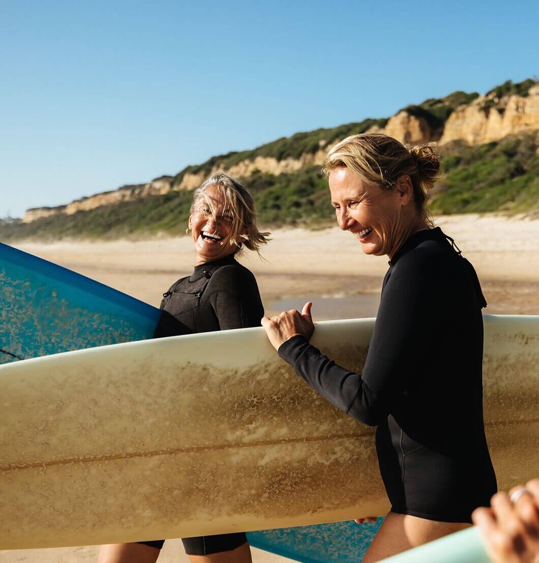 Two older female surfers carrying surfboards and laughing