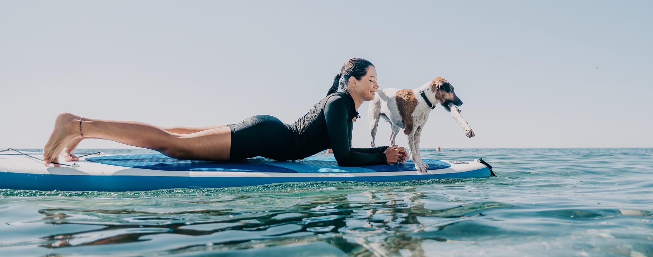 A female surfer laying on a surfboard in the water with a dog next to her
