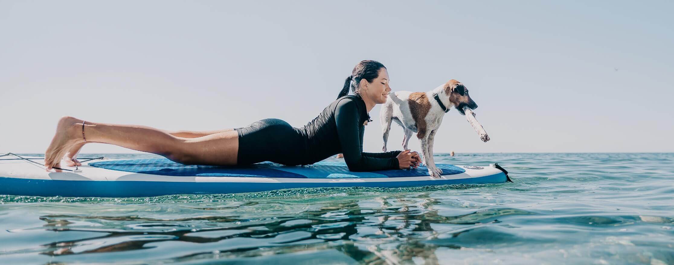 A female surfer laying on a surfboard in the water with a dog next to her
