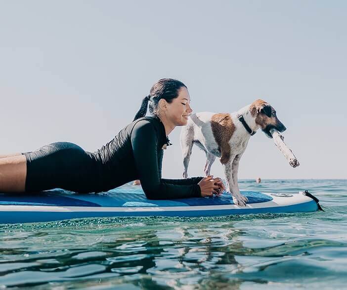 A female surfer laying on a surfboard in the water with a dog next to her